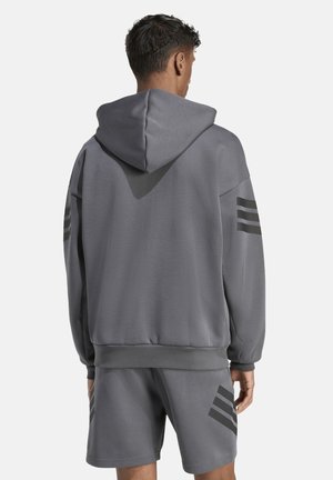 Man draagt een grijze hoodie en bijpassende shorts met zwarte diagonale strepen, staat met zijn rug naar de camera tegen een eenvoudige lichte achtergrond.