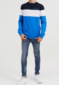 Sudadera de manga larga azul con rayas horizontales en marino, blanco y azul claro. Tiene un cuello redondo y está hecha de un tejido suave. Combinada con jeans ajustados y zapatillas blancas.