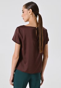 Bordeauxfarbene Seidenbluse mit kurzen Ärmeln, einem subtilen Rückenschlitz und einer glatten Textur, kombiniert mit türkisfarbenen Hosen.