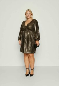 Nő fényes arany színű, hosszú puffos ujjú, kötényruhába öltözve, fekete bokapántos magassarkú cipőben, és egy kicsi fekete clutch táskával a kezében.