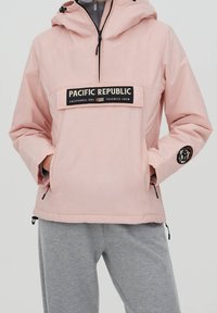 Lyserød vindjakke med hætte, der har en frontlomme og "PACIFIC REPUBLIC" logo. Strukturerede grå sweatpants nedenfor.