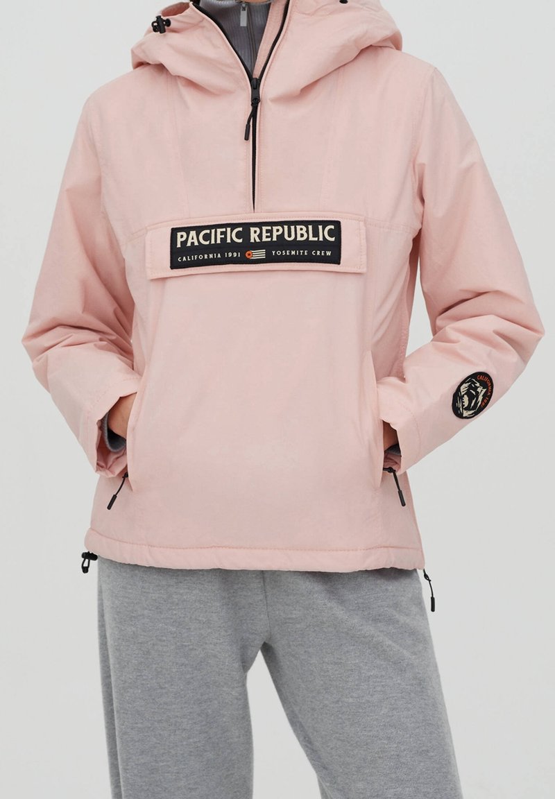 Lyserød vindjakke med hætte, der har en frontlomme og "PACIFIC REPUBLIC" logo. Strukturerede grå sweatpants nedenfor.