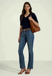 Mulher vestindo uma blusa azul-marinho de decote em V, jeans azuis, cinto castanho, sandálias de salto castanhas e segurando uma bolsa de ombro de couro castanha.