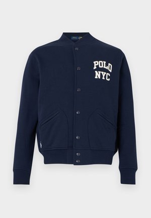 Tumši zila varsity stila jaciņa ar pogām, ribota auduma aprocēm un apakšdaļu, priekšējām kabatām un baltu uzrakstu "POLO NYC" uz krūtīm.