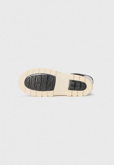 Semelle d'une chaussure noire et crème avec des zones de grip noir texturé et une semelle épaisse crème sur un fond clair.