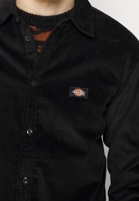 Camicia in velluto a coste nera con chiusura a bottoni, caratterizzata da una piccola toppa con il logo colorato "Dickies" sul petto, tessuto a coste con texture.
