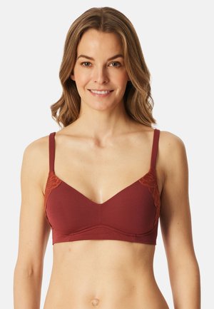 Kvinde iført en rustfarvet bh med blonde detaljer på skålene, smilende, med skulderlangt, bølget hår foran en enkel hvid baggrund.