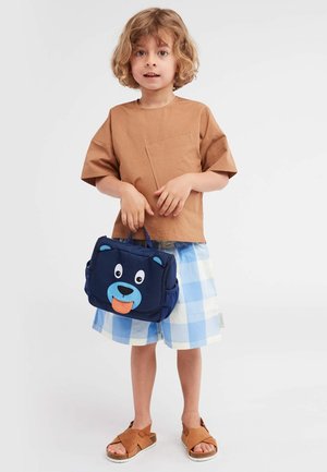 Braunes Baumwollhemd mit kurzen Ärmeln, blaue karierten Shorts, braune Sandalen und eine marineblaue Tasche mit einem Cartoon-Bärenkopf und einem Zungendetail.