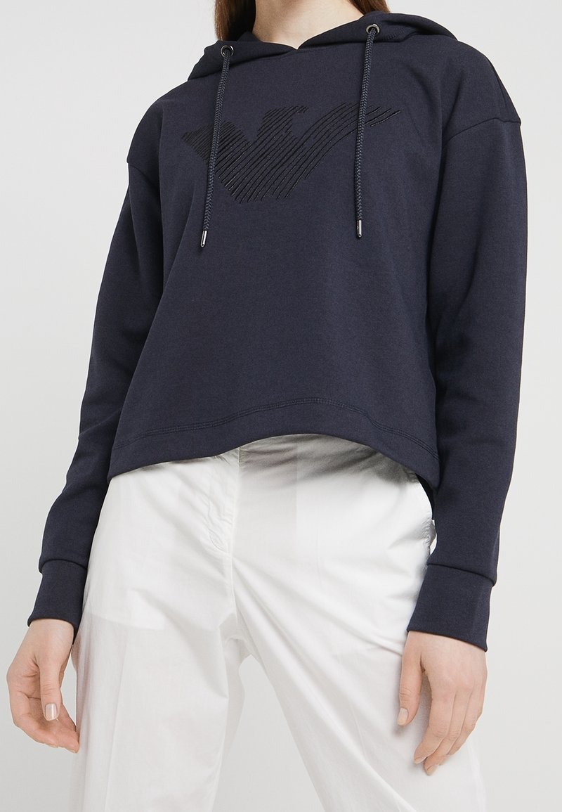 Marinblå kort hoodie i mjukt tyg, med en texturerad logodesign på bröstet och dragsnöre i huvan. Vita byxor nedanför.