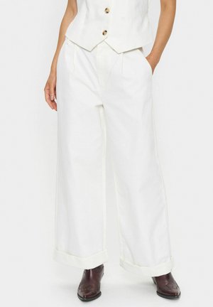 Pantaloni bianchi a gamba larga con pieghe frontali, caratterizzati da un risvolto e cuciture a contrasto color crema, abbinati a stivali marroni.
