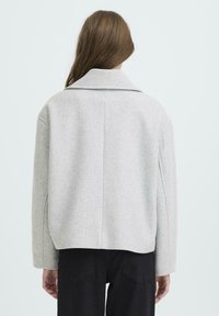 Femme portant une veste en laine grise claire avec un large col et des manches longues, associée à un pantalon noir, se tenant de dos.