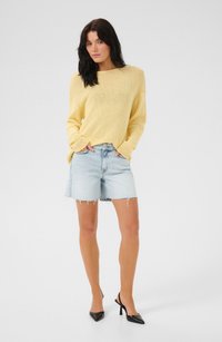 Suéter amarillo de punto con un corte holgado, combinado con pantalones cortos de denim azul claro con los bordes deshilachados y zapatos de tacón alto negros tipo slingback.