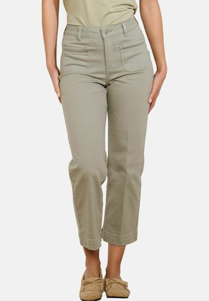 Femme portant un pantalon beige taille haute, raccourci, avec des poches avant et des mocassins beiges, debout devant un fond blanc.