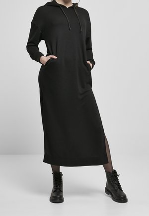 Musta hupullinen midimekko, jossa on pitkät hihat, taskut ja rento leikkaus. Valmistettu pehmeästä kankaasta, jossa on sileä tekstuuri, ja yhdistetty mustiin nauhallisiin saappaisiin.