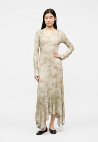 Γυναίκα που φοράει μπεζ μακρυμάνικο φόρεμα με διακριτικό λουλουδάτο μοτίβο και ασύμμετρο τελείωμα, συνδυασμένο με μαύρα επίπεδα παπούτσια, στέκεται με το χέρι στη μέση.