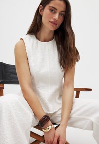 Hvid struktureret to-delt sæt bestående af en ærmeløs top og brede bukser. Accessories inkluderer et mørkt armbånd og et guldarmbånd.