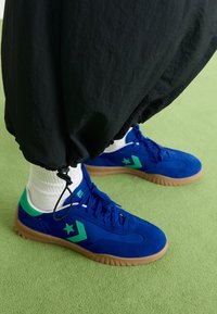 Des baskets en daim bleu avec des accents verts, un logo étoile et une semelle en caoutchouc gomme texturée. Portées avec des chaussettes blanches et un pantalon noir.