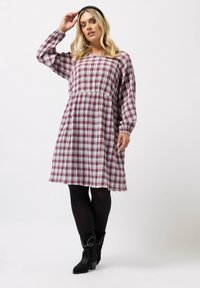 Nainen, joka on pukeutunut löysään, pitkähiharaiseen vaaleanpunaisiin ja valkoisiin ruutukuvioisiin mekkoihin, mustiin leggingseihin, mustiin nilkkabootseihin ja mustaan hiuspantaan valkoista taustaa vasten.