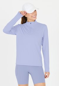 Kvinde smilende, iført hvid baseballkasket, lyse lilla langærmet lynlåstrøje og matchende cykelshorts med høj talje, stående mod en hvid baggrund.