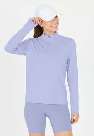 Kvinna som ler, bär vit basebollkeps, ljuslila långärmad zip-up tröja och matchande högmidjade cykelshorts, stående mot vit bakgrund.