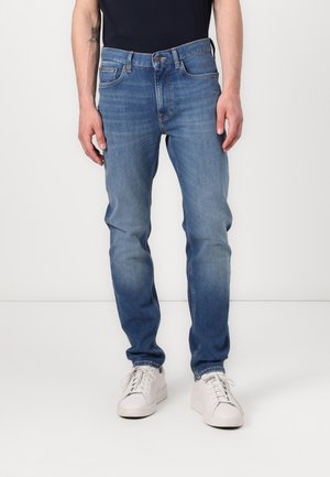 Mand iført slim fit blå jeans og hvide sneakers står mod en ensfarvet baggrund med hænderne afslappet ned langs siderne.