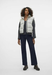 Gilet imbottito grigio con colletto alto, abbinato a un maglione lavorato scuro, jeans denim a gamba larga scuri e stivaletti marroni. 