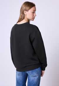 Czarny, oversizowy sweatshirt z okrągłym dekoltem i prążkowanymi mankietami, wykonany z miękkiego, teksturowanego materiału. Noszony z wypłowiałymi niebieskimi dżinsami.