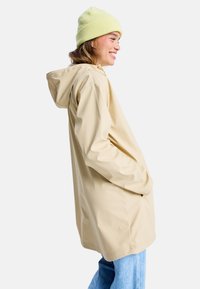 Helle beigefarbene wasserfeste Jacke mit Kapuze, langen Ärmeln und Seitentaschen; getragen mit einer weichen, gelben Strickmütze. Lässiger Schnitt und glatte Textur.