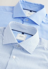 Deux chemises habillées : l'une est unie, bleu clair, avec une finition texturée, et l'autre est blanche avec des rayures bleues. Les deux ont des cols boutonnés et des étiquettes visibles.