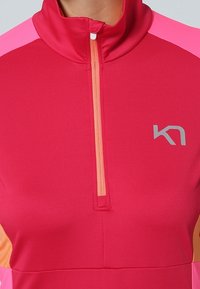 Jasnoczerwona sportowa bluzka z wysokim kołnierzem, z różowymi i pomarańczowymi akcentami. Posiada przedni zamek błyskawiczny i subtelne logo.