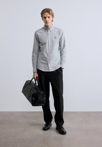 Γκρι πουκάμισο με κουμπιά και μικρό έμβλημα, συνδυασμένο με μαύρο παντελόνι και μαύρη τσάντα duffel με υφή. Τα παπούτσια είναι μαύρα και γυαλισμένα.