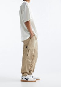 Hvid oversized t-shirt, khaki cargo bukser med lommer, og hvide sneakers med sorte accenter og gummisåler, set fra siden.