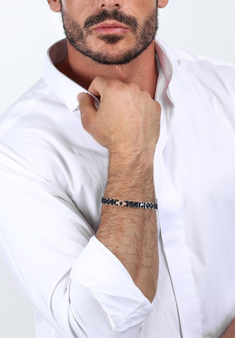 Bracciale in metallo con maglie nere e argento alternate, caratterizzato da un sottile accento con logo, indossato su un polso con una camicia a maniche lunghe di colore chiaro.