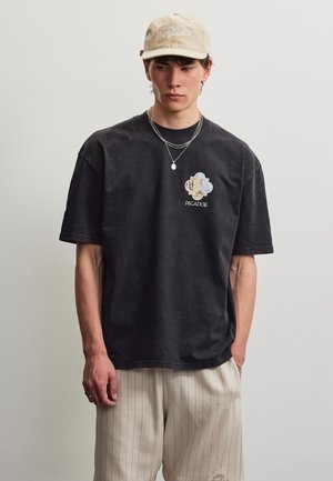 Ung mand iført en beige kasket, sort grafisk t-shirt med "PEGADOR" logo, lag af sølvhalskæder og lyse bukser med striber mod en ensfarvet baggrund.