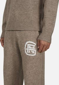 Brązowy sweter dzianinowy i spodnie wykonane z miękkiego materiału. Sweter ma ściągacze u mankietów; spodnie mają na udzie wyszyte białe logo.