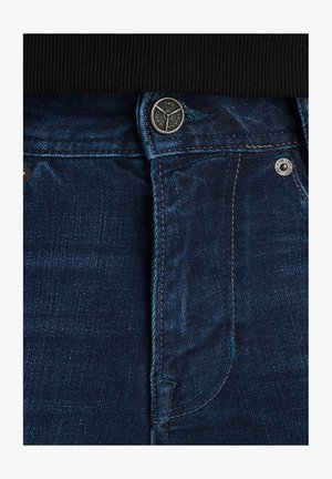 Vaqueros de mezclilla en azul oscuro con una textura suave. Cuenta con un cierre de botón de metal, costuras en contraste y una cinturilla visible en la parte superior.