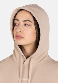 Sudadera de algodón beige con cordones, que presenta un bolsillo frontal tipo canguro. La tela es suave y tiene un ligero brillo. Logo impreso en blanco.