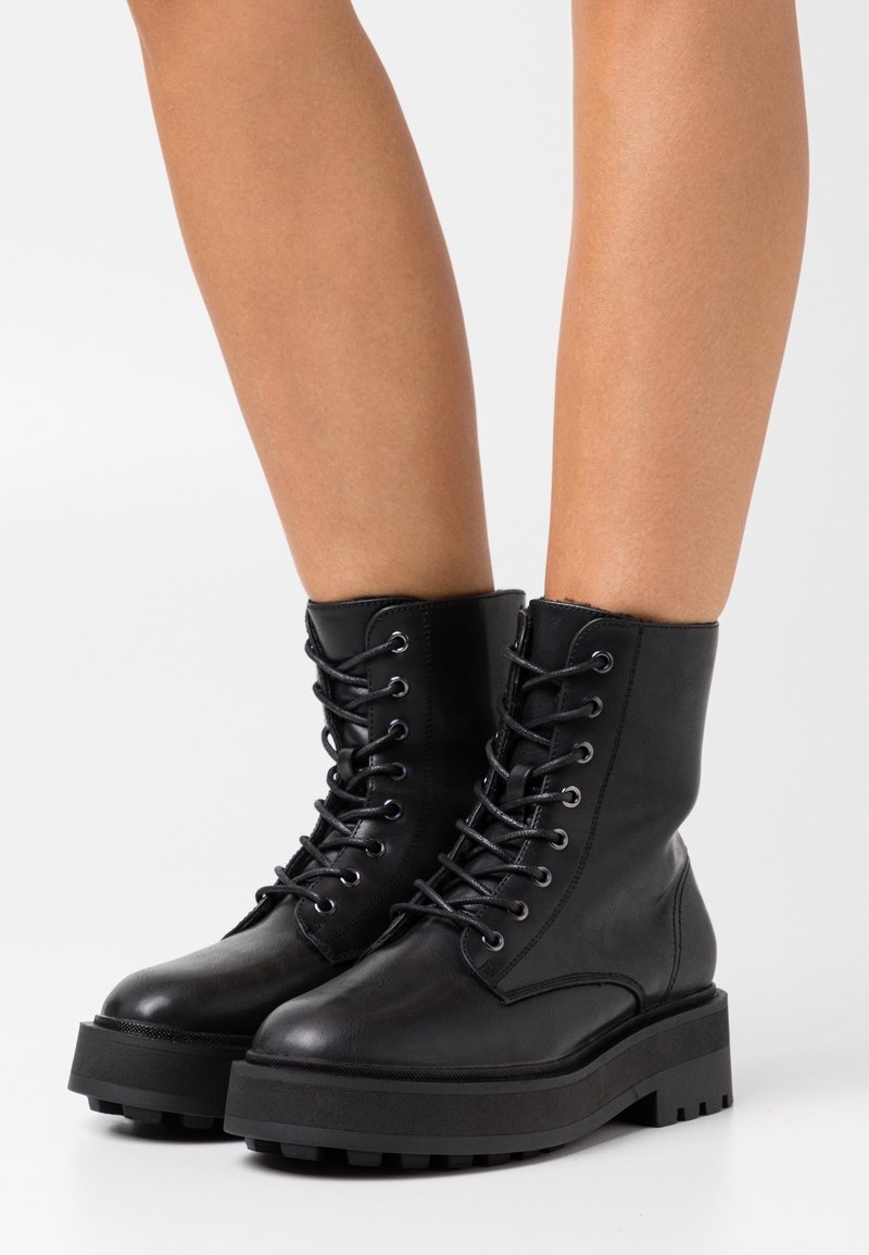 Botas de combate de cuero negro con suela gruesa negra, cordones en la parte delantera, textura suave y ojales reforzados. Altura hasta el tobillo con un diseño robusto.