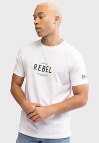 Hvid bomulds-T-shirt med sort "REBEL" tryk på fronten og venstre ærme. Korte ærmer og rund halsudskæring, afslappet pasform.