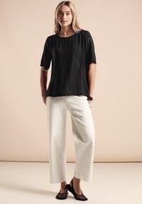 Top corto nero a maniche corte con linee astratte bianche, abbinato a pantaloni larghi bianchi e scarpe aperte nere con un motivo testurizzato.