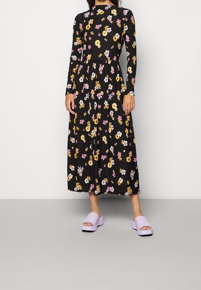 Femme portant une robe midi noire à manches longues avec des motifs floraux jaunes, blancs et roses et des sandales plateformes lilas.