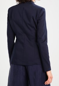 Námořnický upravený blazer, strukturovaný design, hladká tkanina, jediný zadní otvor, přiléhavý silueta, dlouhé rukávy, minimální detaily prošívání.