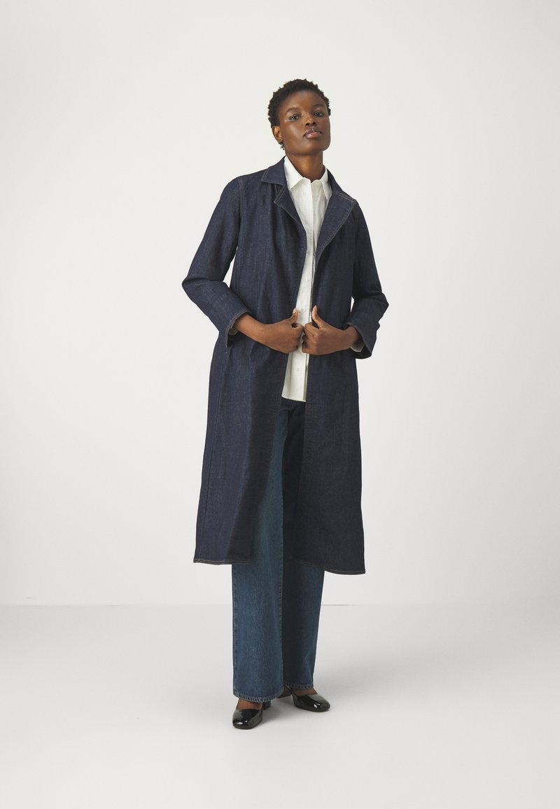 Cappotto lungo in denim scuro con colletto, apertura frontale e maniche arrotolate, indossato sopra una camicia bianca e pantaloni larghi in denim scuro. Scarpe nere.