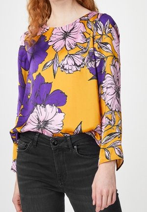 Vrouw draagt een mosterdkleurige blouse met lange mouwen en grote paarse en roze bloemenpatronen, gecombineerd met zwarte high-waist jeans.