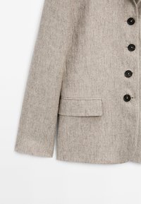 Blazer gris clair en tissu texturé. Il comporte une poche poitrine unique, trois boutons noirs sur le devant et une coupe cintrée.