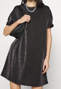 Robe noire à manches courtes avec finition métallique, coupe ample et manches roulées. Accompagnée d'un sac à main noir et d'un collier en chaîne argenté.