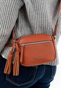 Borsa a tracolla in pelle marrone con finitura testurizzata, dotata di tasca frontale con zip e due dettagli a frange. Indossata insieme a un maglione grigio.