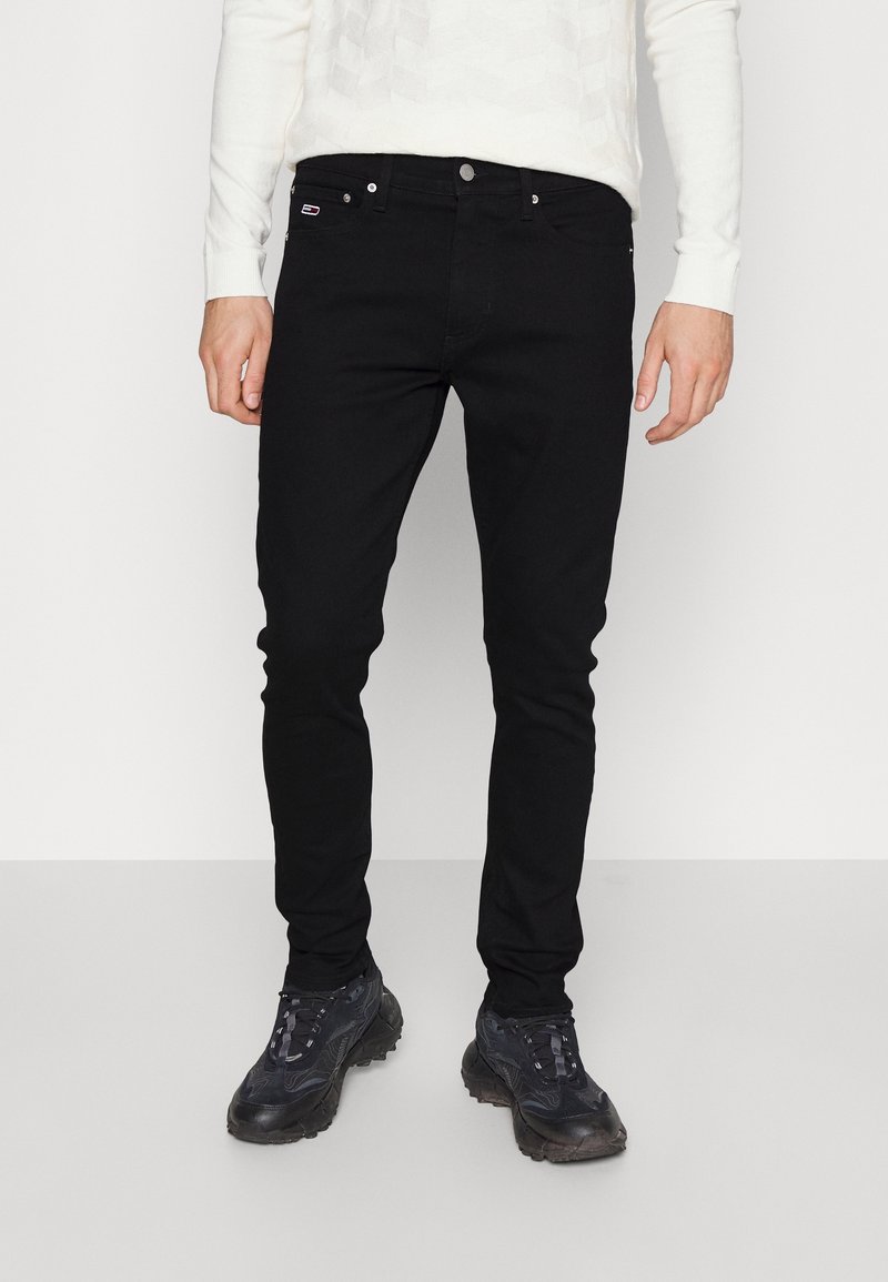 Man wearing slim black jeans, white long-sleeve shirt, and black athletic shoes standing against a plain background.