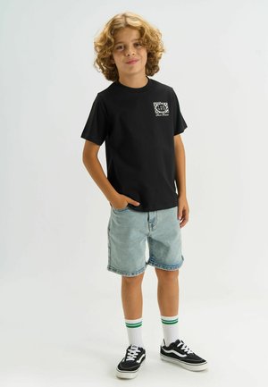 Chłopiec z kręconymi włosami, ubrany w czarny t-shirt z grafiką, jasne jeansowe szorty, białe skarpetki z zielonymi paskami i czarne tenisówki, stoi z ręką w kieszeni.