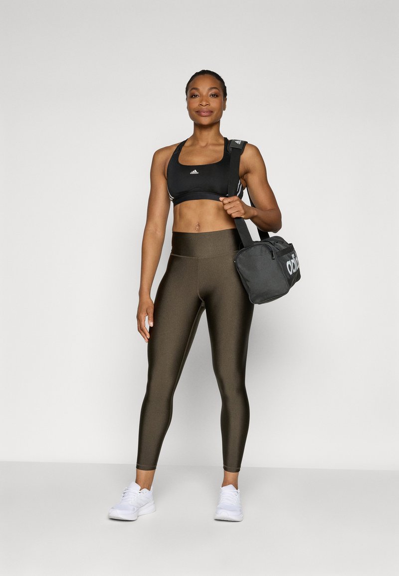 Schwarzer Sport-BH, olivgrüne hochtaillierte Leggings und weiße Sportschuhe. Hält eine schwarze Sporttasche mit weißer Beschriftung.